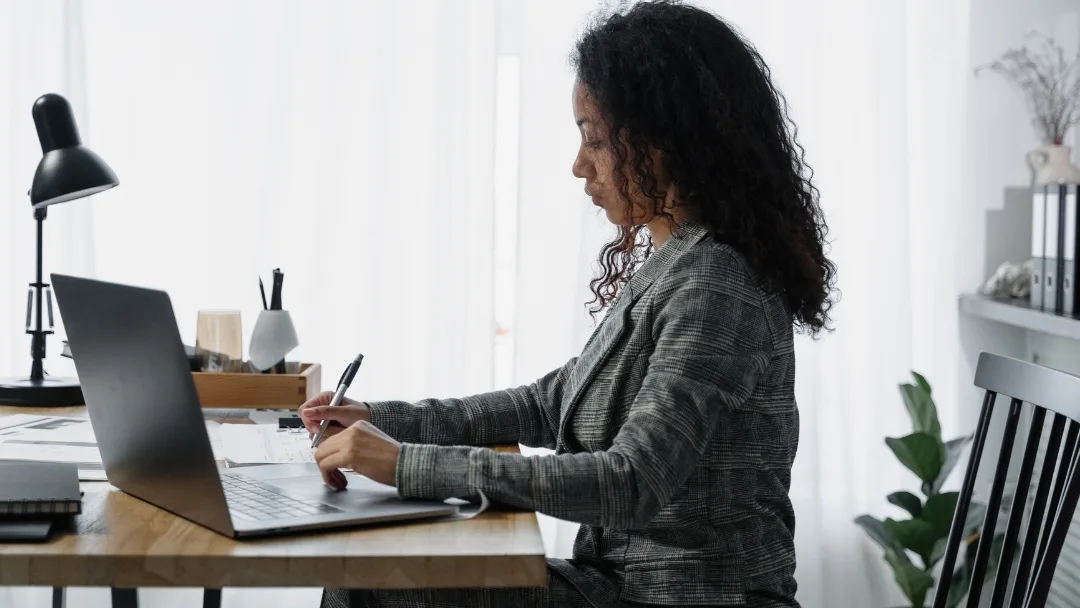 A woman holding a pen and looking at her budgeting plan on her laptop while managing her chronic illness.